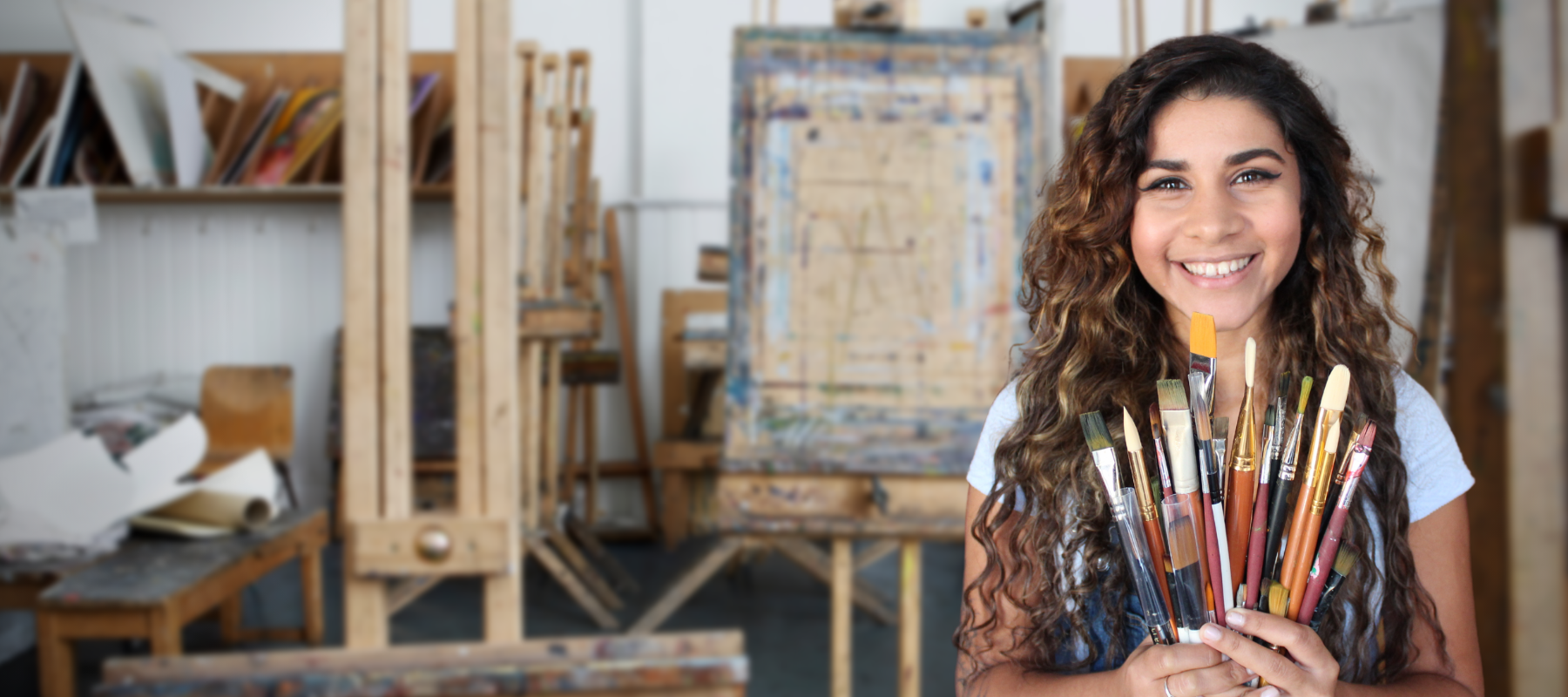 artist Sema Martin, woman with curly brown and blond hair holding a hand full of paint brushes with art easels behind her